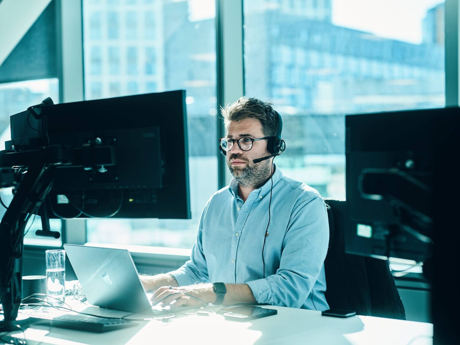 Man wearing a headset sits at a desk with a laptop and two monitors in a bright office.