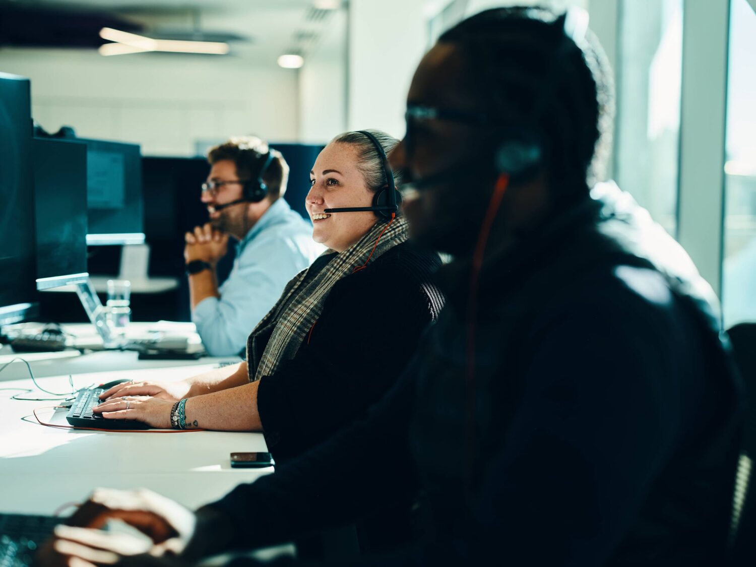 Customer service agents wearing headsets work at computer desks in a bright office.