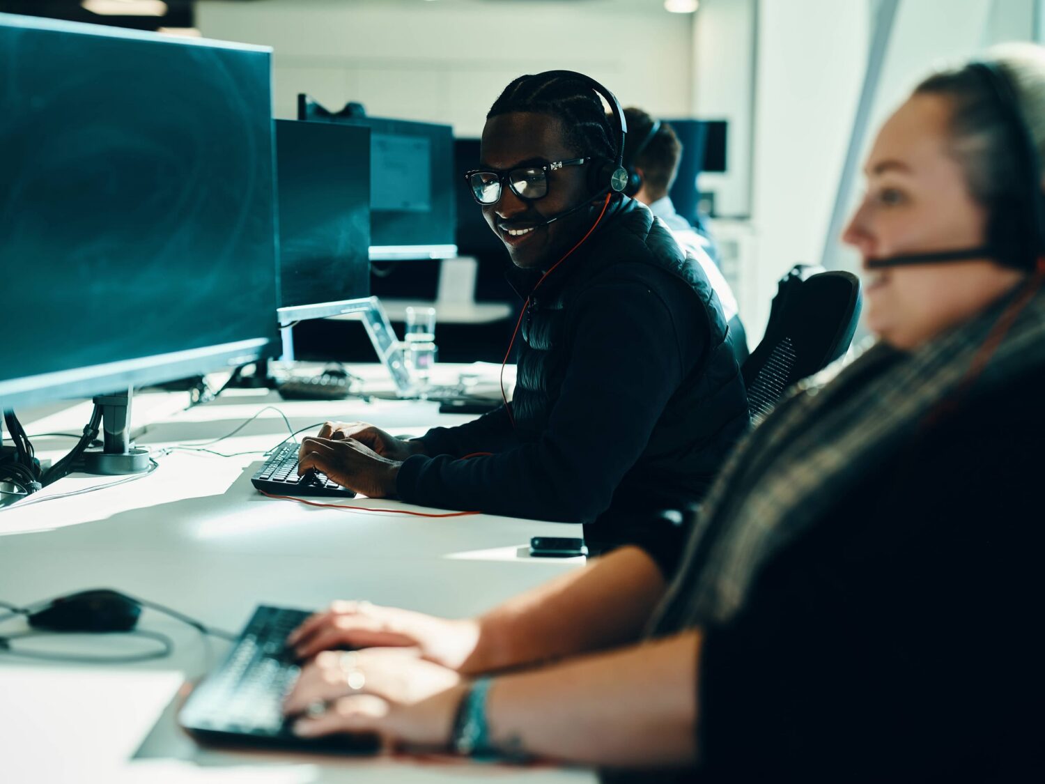 Two colleagues wearing headsets work at computer desks in a bright, modern office.