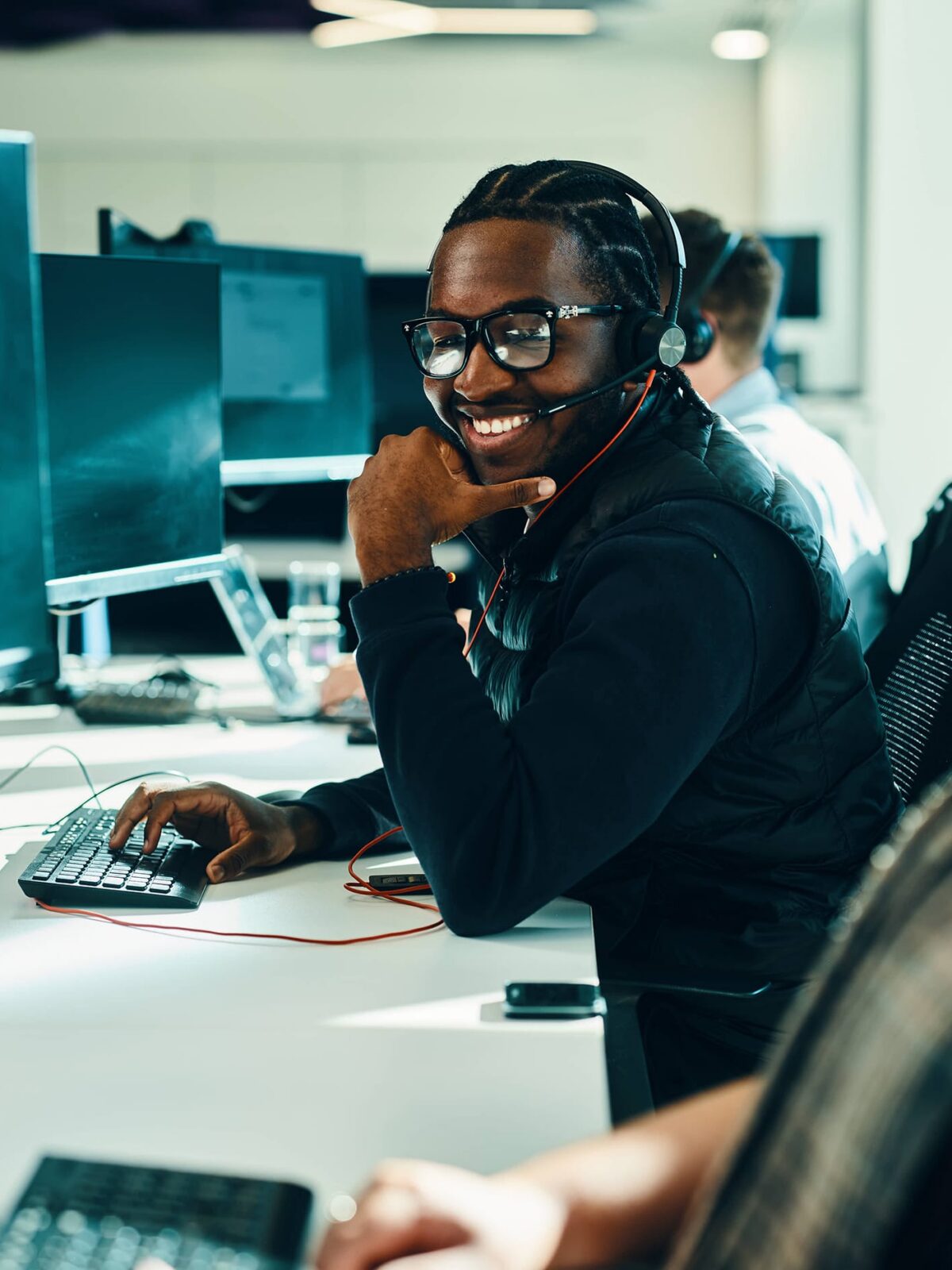 A smiling customer service representative wearing a headset sits at a desk with a computer in a busy call center.