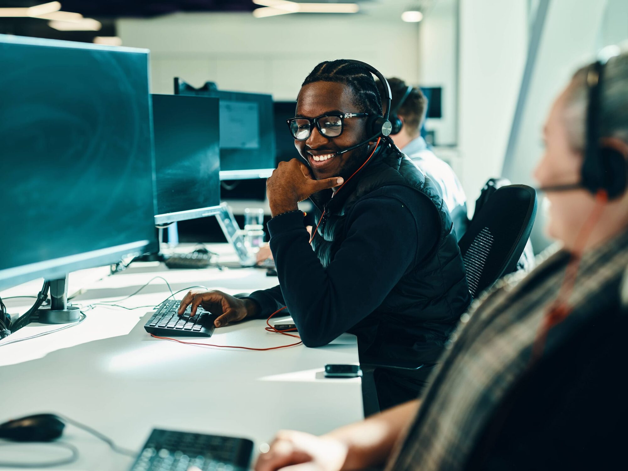 A smiling customer service representative wearing a headset sits at a desk with a computer in a busy call center.