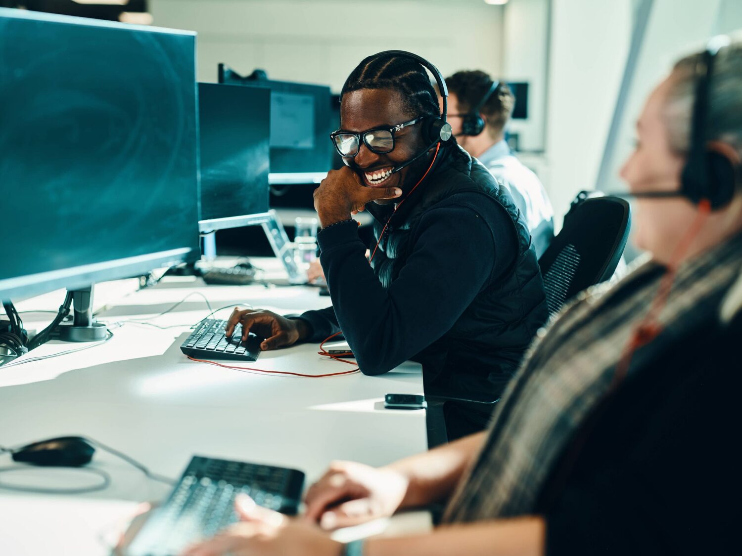 A smiling woman wearing a headset sits at a computer in a bright call-center office, with colleagues working at desks in the background.