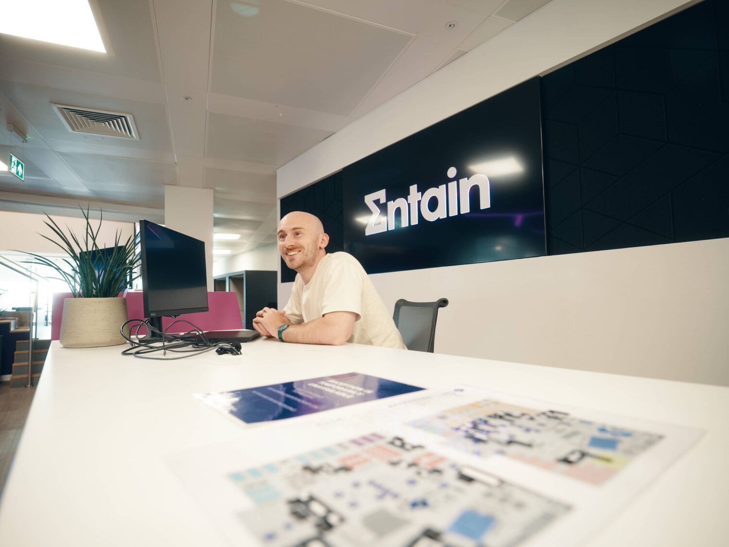 A bald man sits at a white reception desk in a modern office, with a company logo on the dark wall behind him.