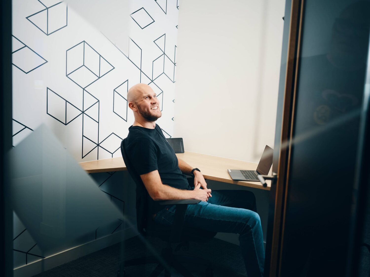 Bearded man in a black T-shirt and jeans sits in a chair at a desk in a modern office with a geometric cube-pattern wall and a laptop.
