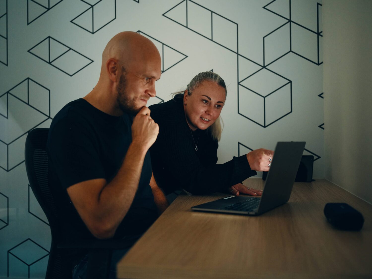 Two people sit at a wooden desk with a laptop, the woman pointing at the screen while the man contemplates.
