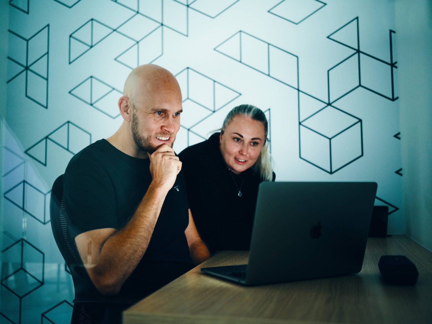 A man and a woman sit at a desk using a laptop, against a wall with geometric cube patterns.
