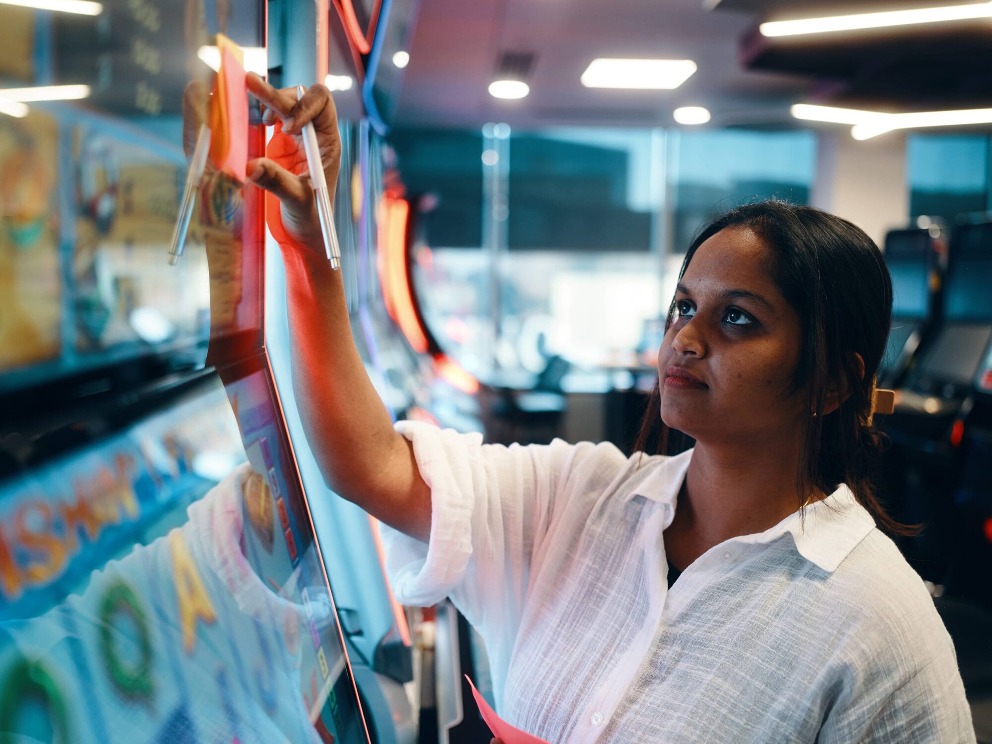 A woman in a white shirt taps a colorful touchscreen on a vending machine in a brightly lit indoor space.