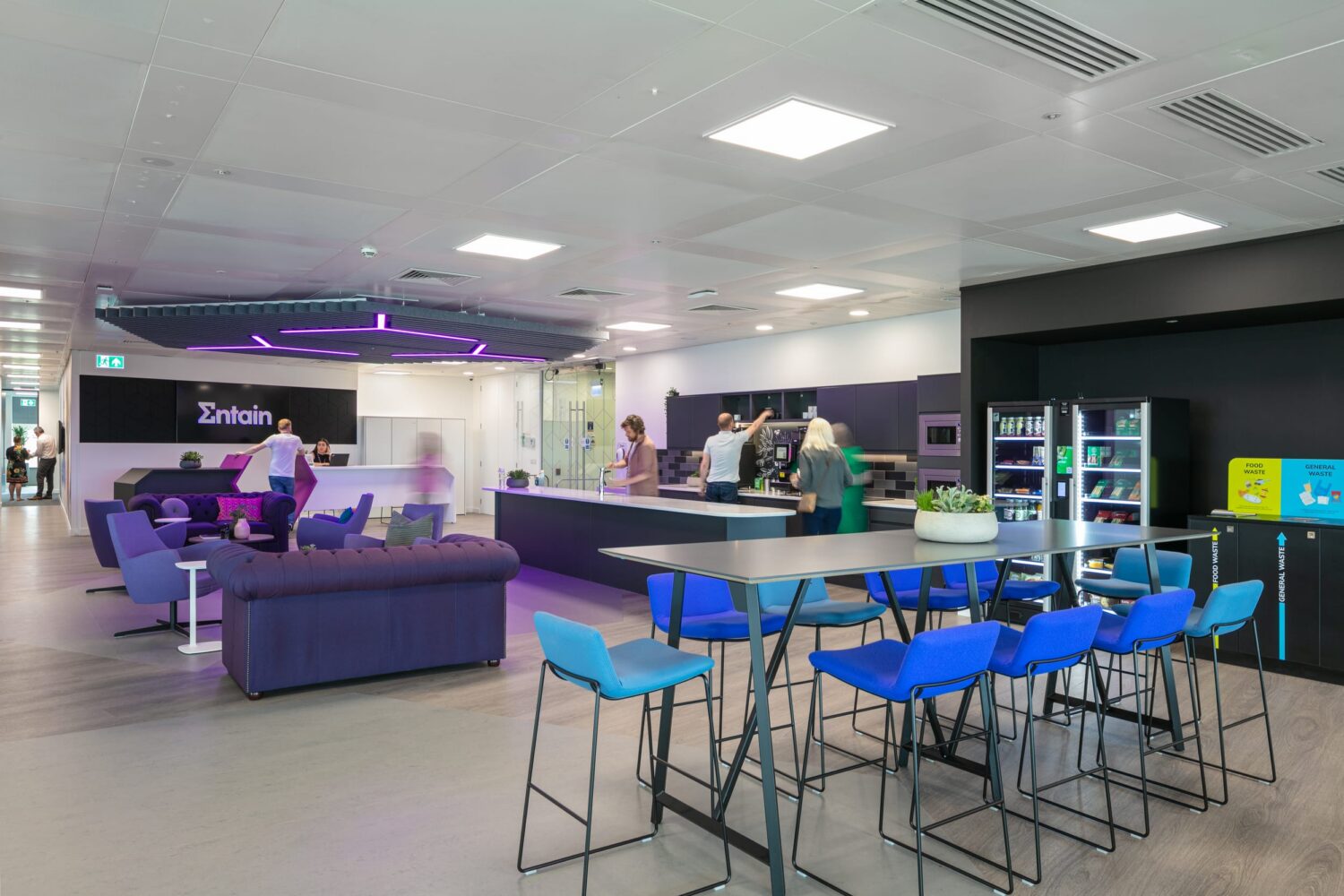 Bright, modern indoor lounge area with purple couches, blue bar stools around a tall high table, and a service counter in the background.