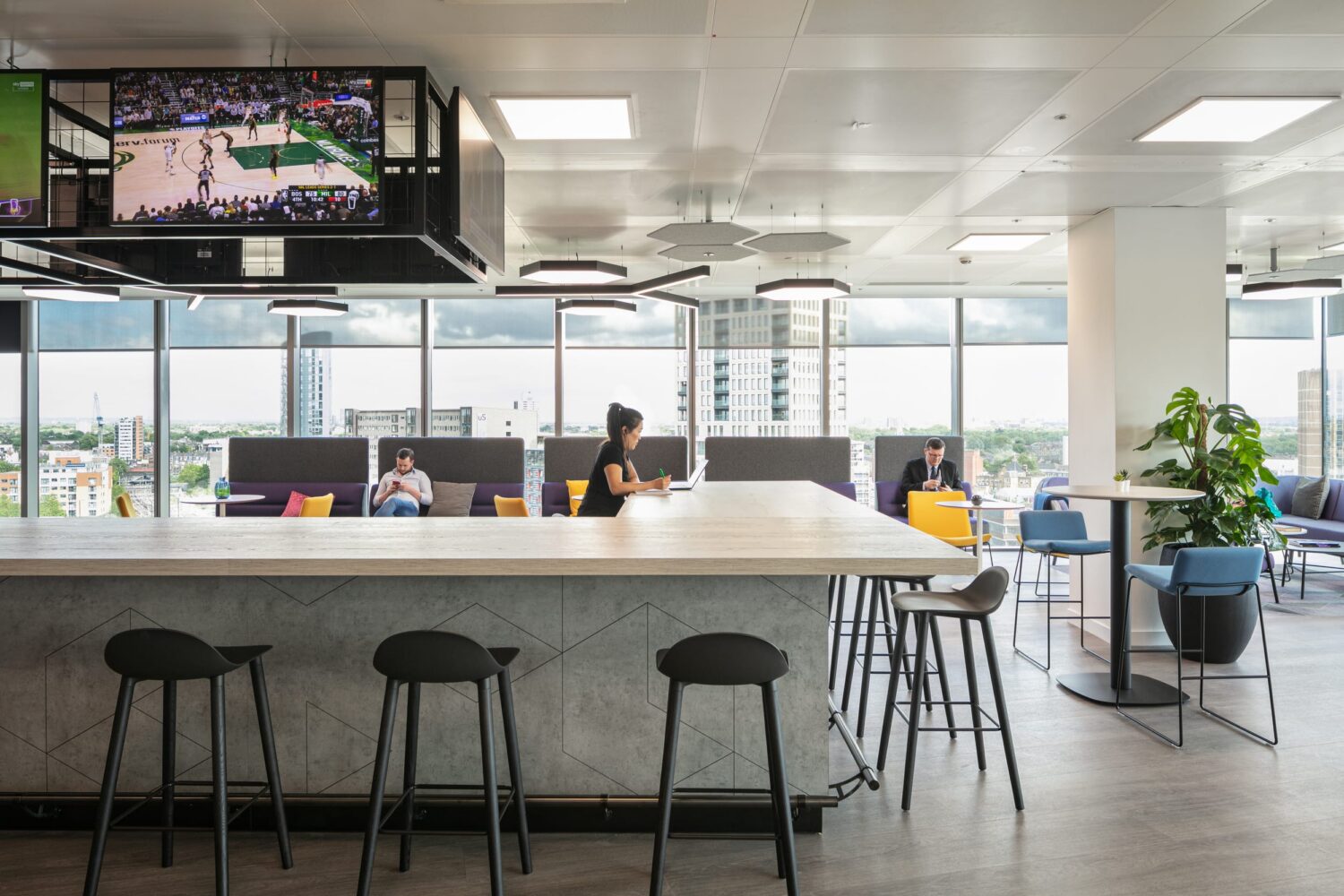 A modern coworking space with a long white counter, black bar stools, and people working by large windows.