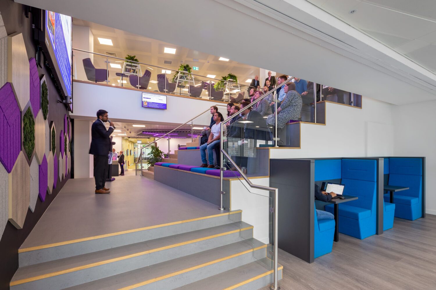 Contemporary indoor public space with a glass-railed staircase, blue seating pods, and people on a mezzanine beside a purple-patterned wall.