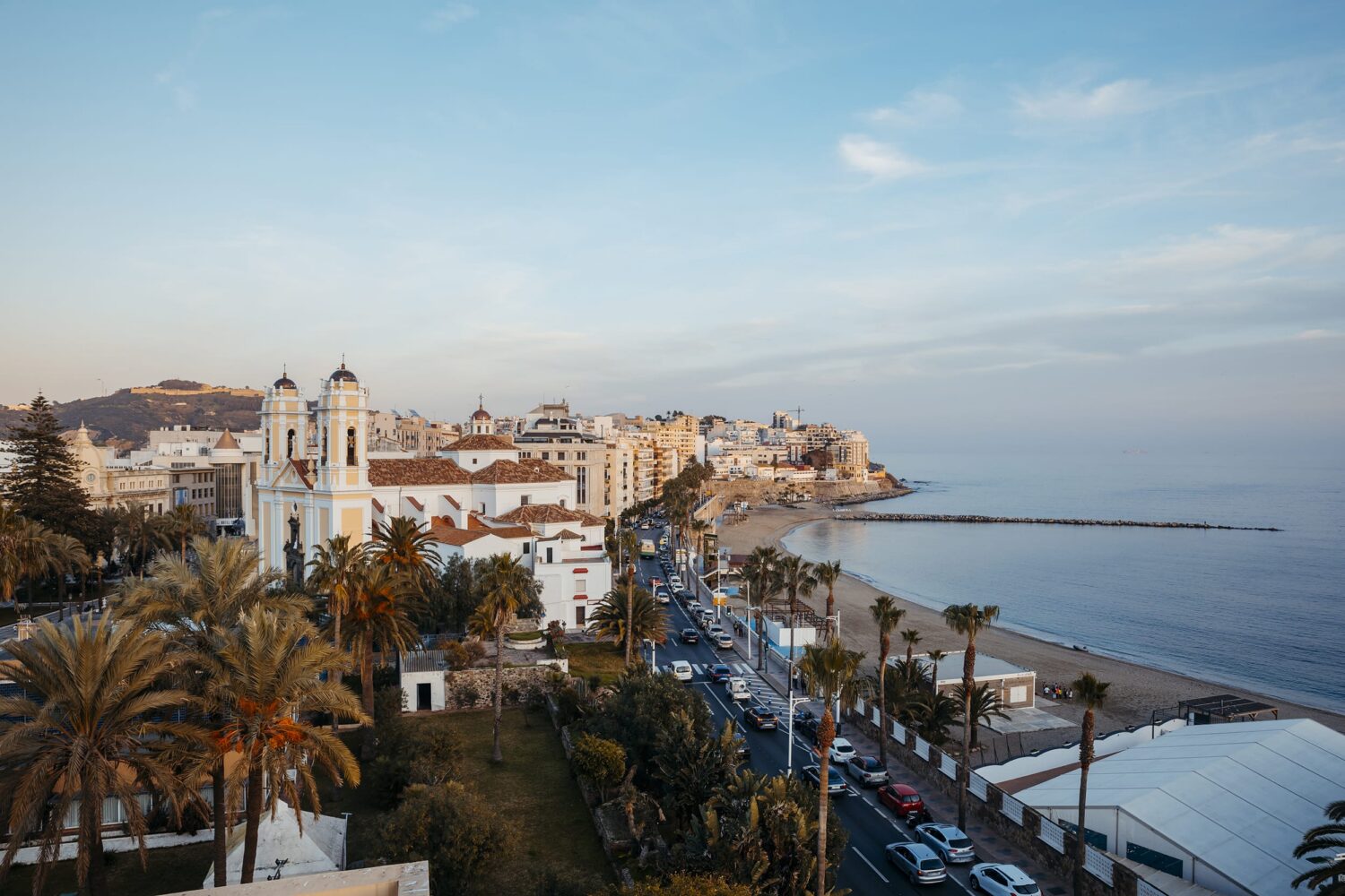 Palm-lined promenade along a calm blue sea, with white and pastel buildings and a rocky headland in the distance.
