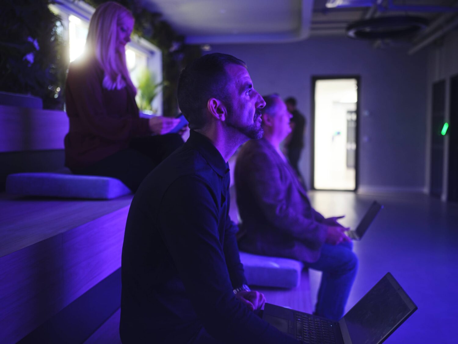 People sit in a dim purple-lit room watching a screen, with a man in the foreground using a laptop.