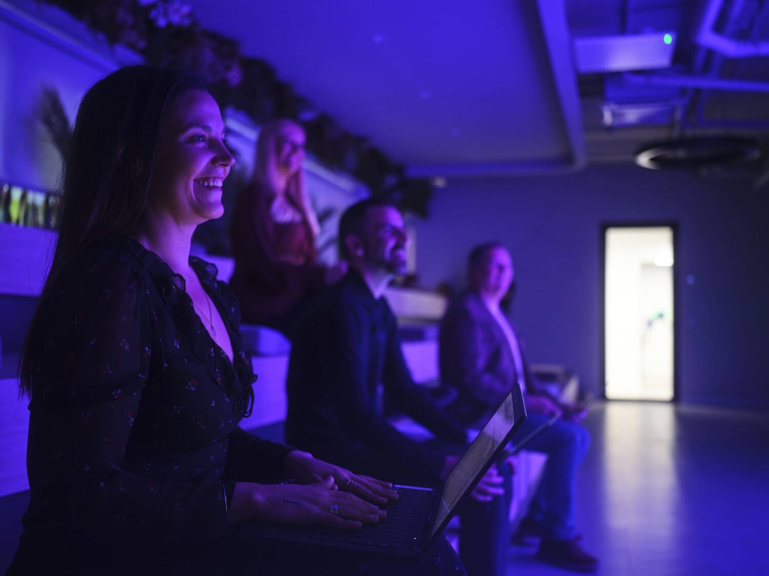 A group of people sit in a dim, purple-lit room, with a smiling woman in the foreground holding a laptop.