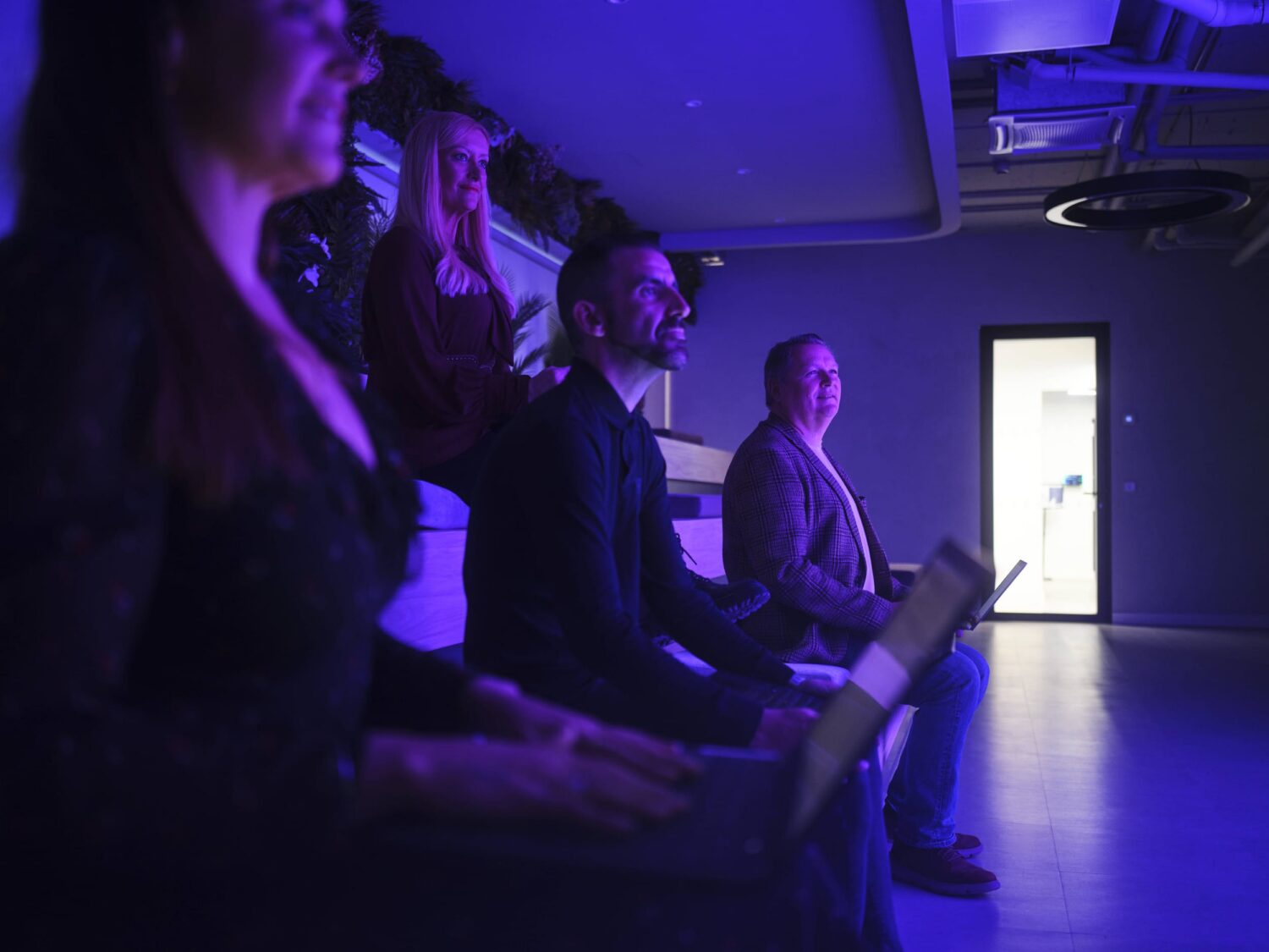 A group of people seated in a dark, purple-lit room watching a presentation, with a bright doorway at the back.
