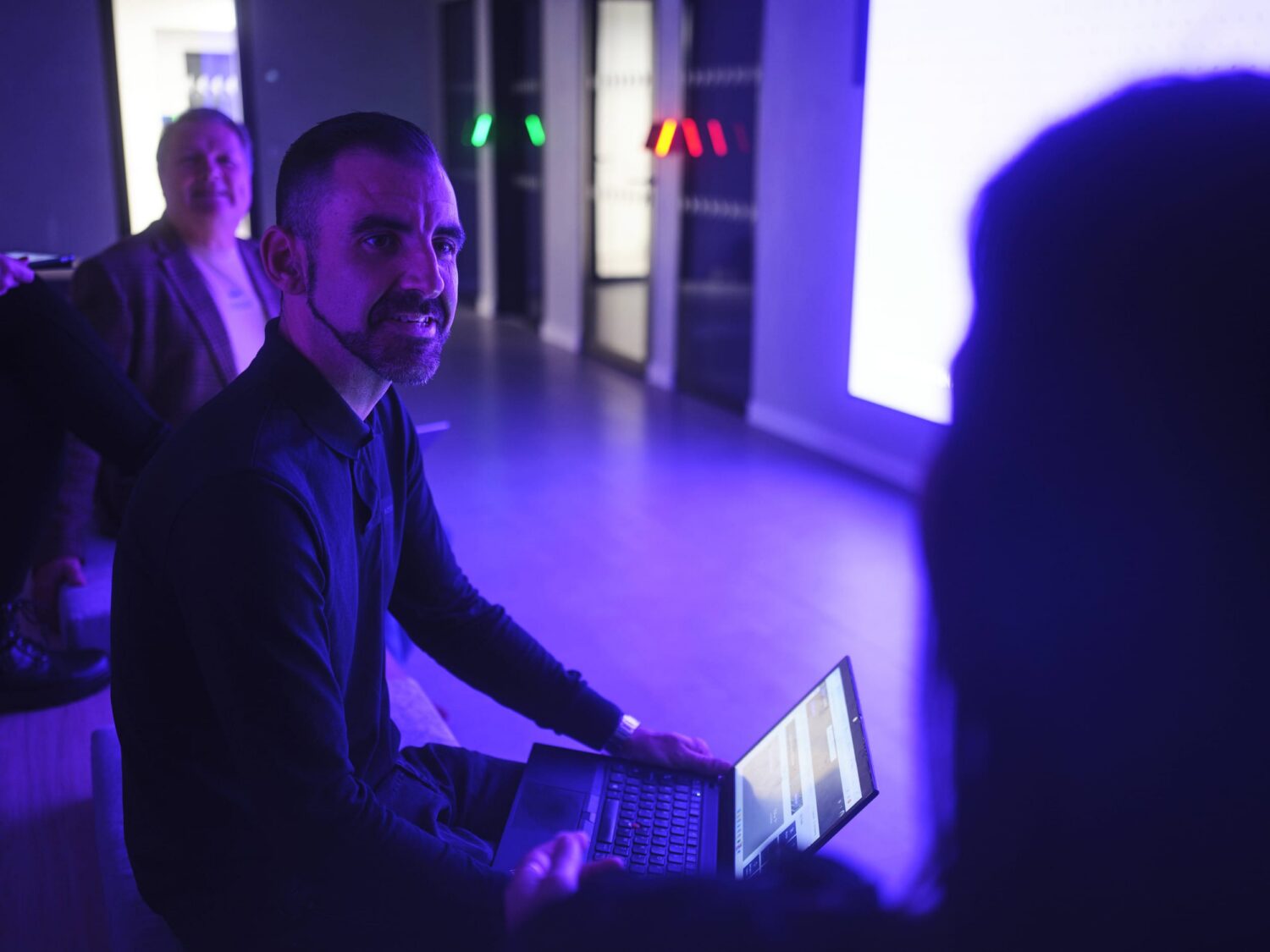 Bearded man sitting with a laptop in a blue-lit event space, looking at the camera.