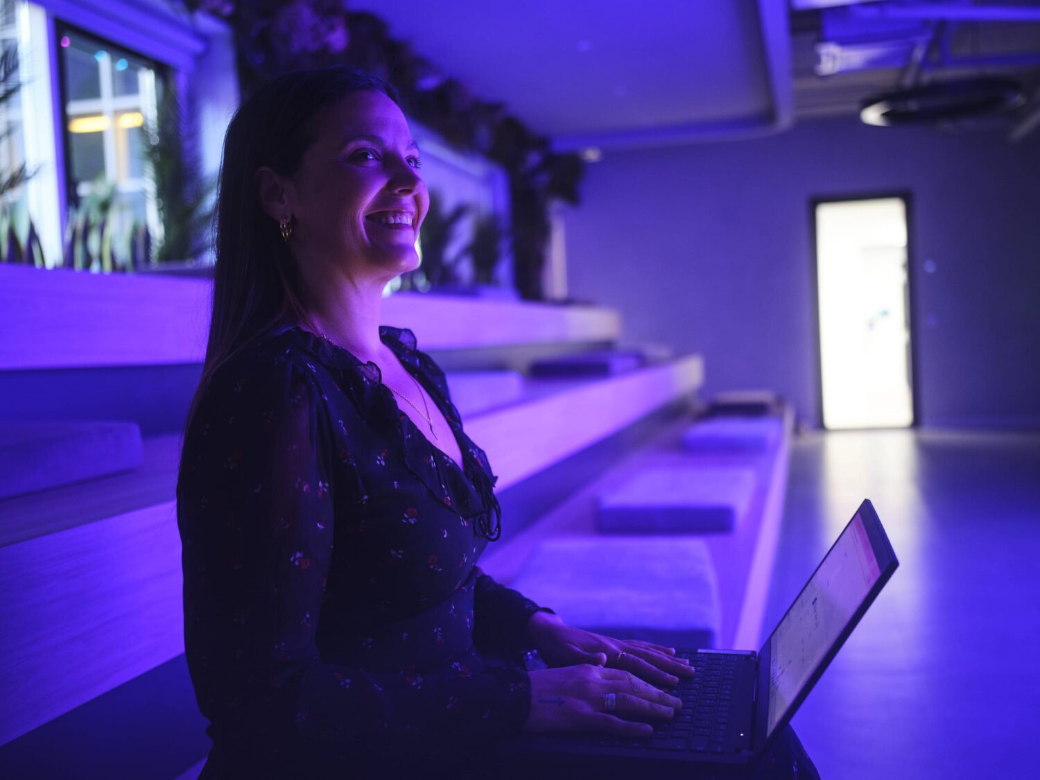 Smiling woman sitting with a laptop on her lap in a blue-lit room with tiered seating.