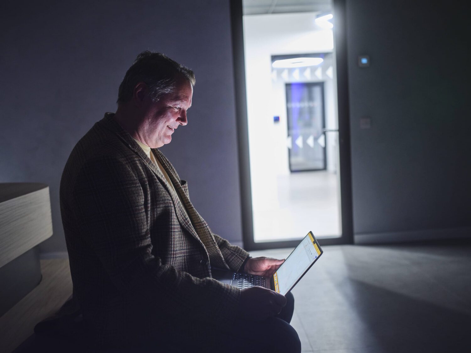 An older man sits in a dim room, illuminated by a tablet he is using, with daylight streaming in from an open door in the background.