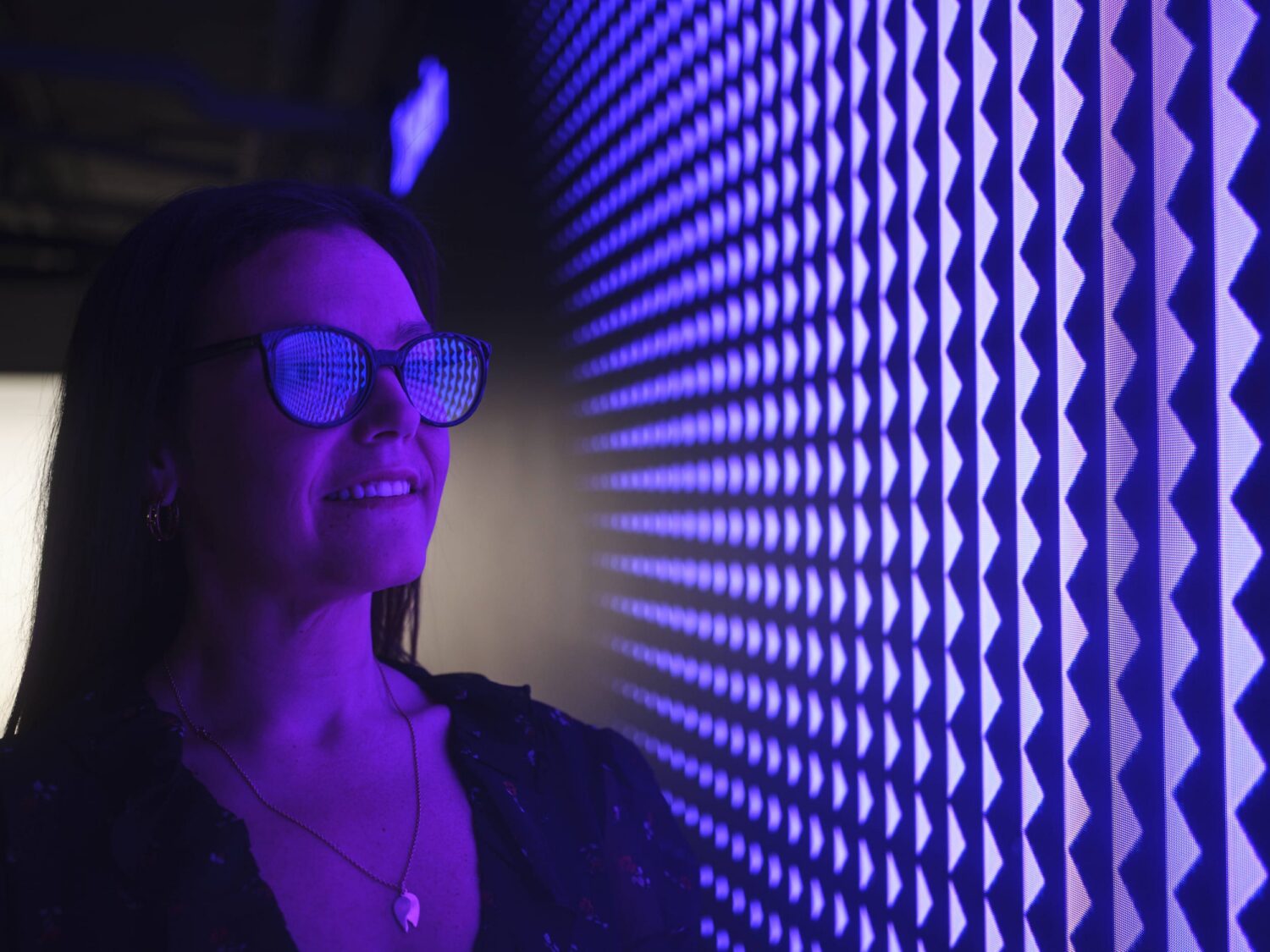 Woman with glasses smiles beside a purple-lit wall featuring a repeating diamond pattern.