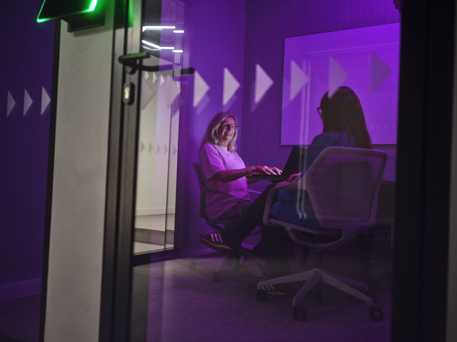 Two people sit and talk inside a purple-lit glass-walled room.