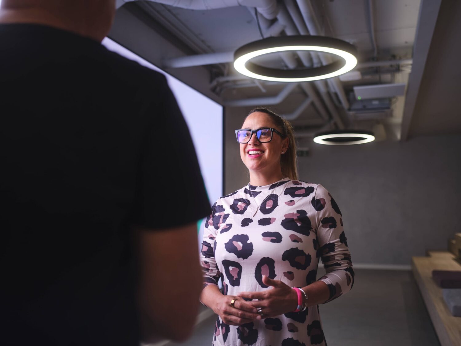 A smiling woman wearing a leopard print dress and glasses talks to a person in a modern room with circular pendant lights.
