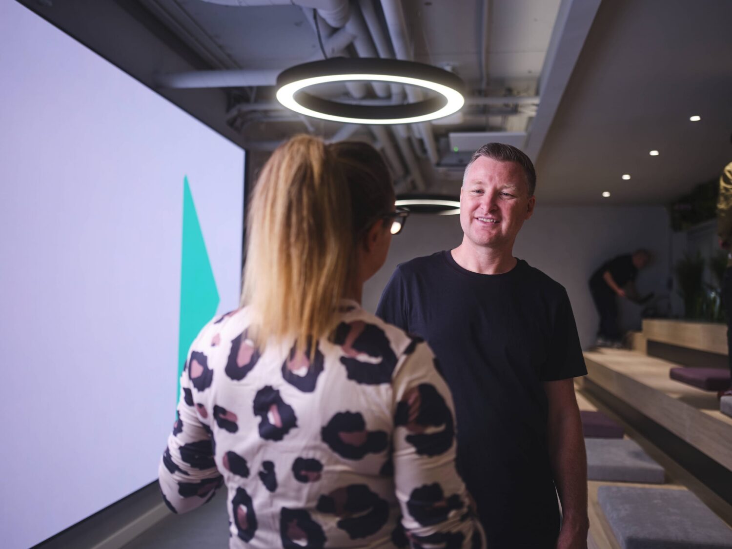 A man smiles at the camera while a woman in a leopard-print sweater stands with her back to the viewer in a modern studio with a circular ring light overhead.