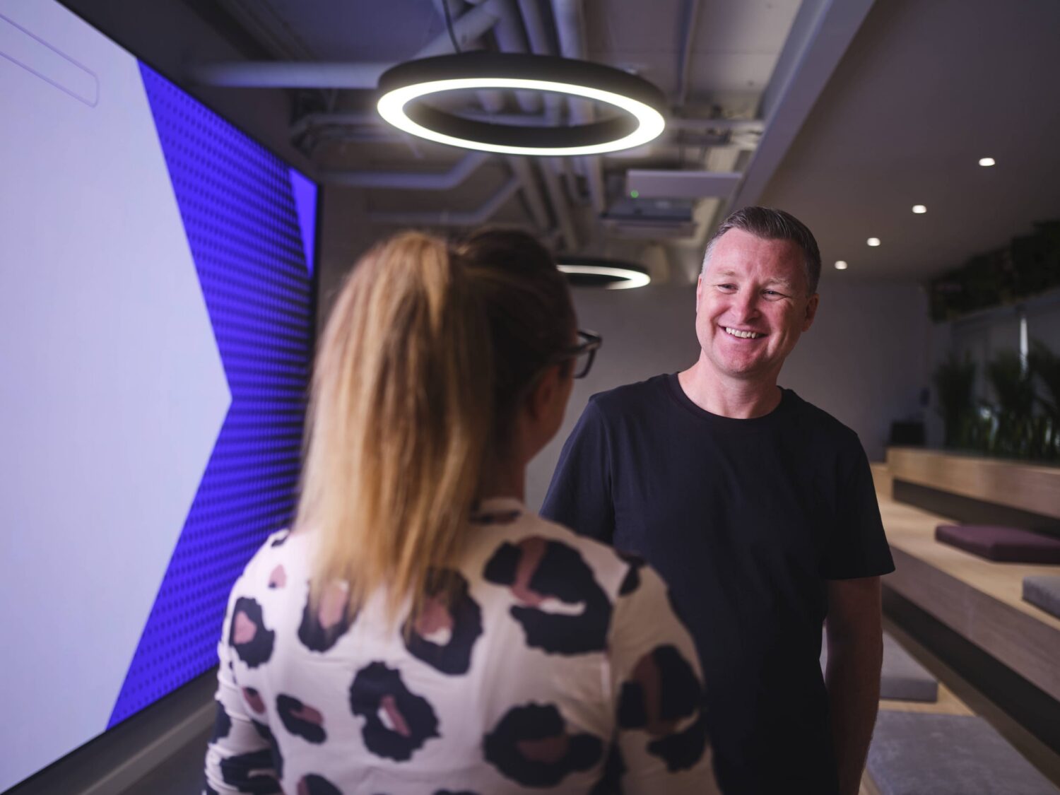A man smiles at a woman in a modern studio with circular ceiling lights and a purple wall.