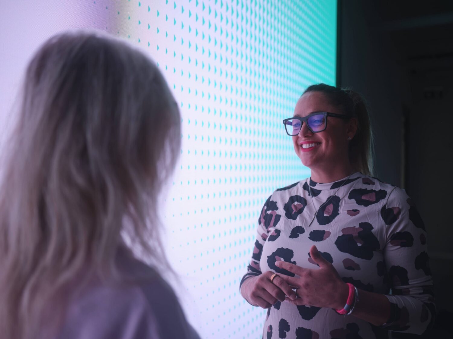 Two people stand near a bright LED wall, with a smiling woman wearing glasses and a patterned sweater talking to the other person.