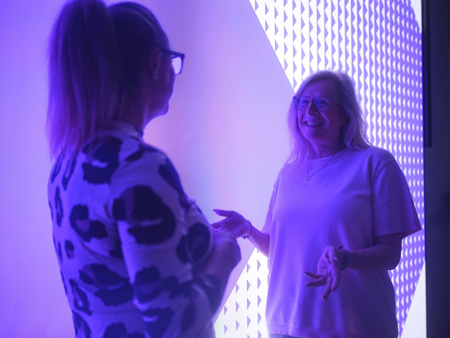 Two women chat in a purple-lit room beside a wall of illuminated LED panels, one with a ponytail and glasses wearing a patterned top, the other in a light T‑shirt, both smiling.
