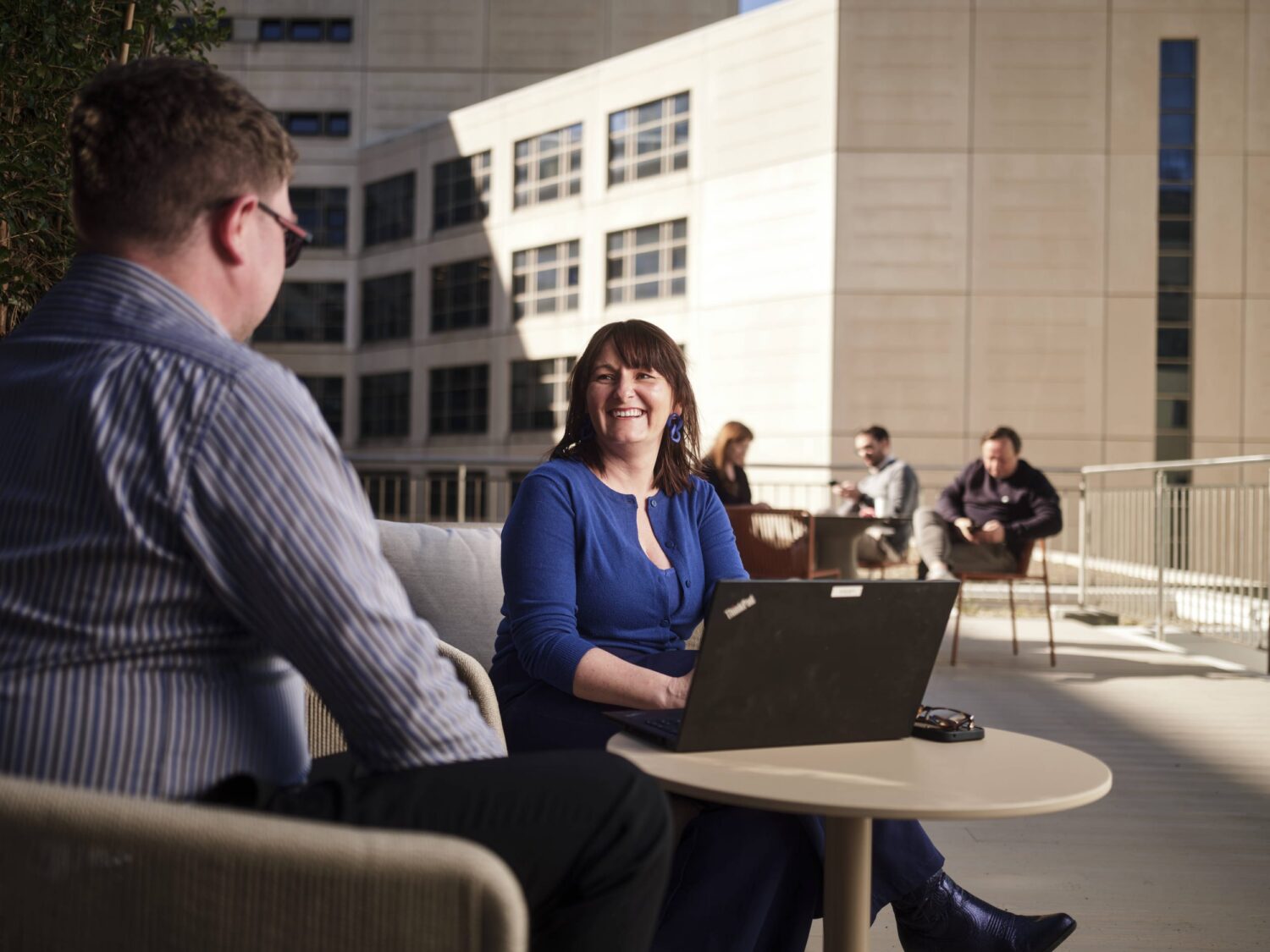Two people sit at a small round table on an outdoor patio, with a laptop on the table and office buildings in the background.