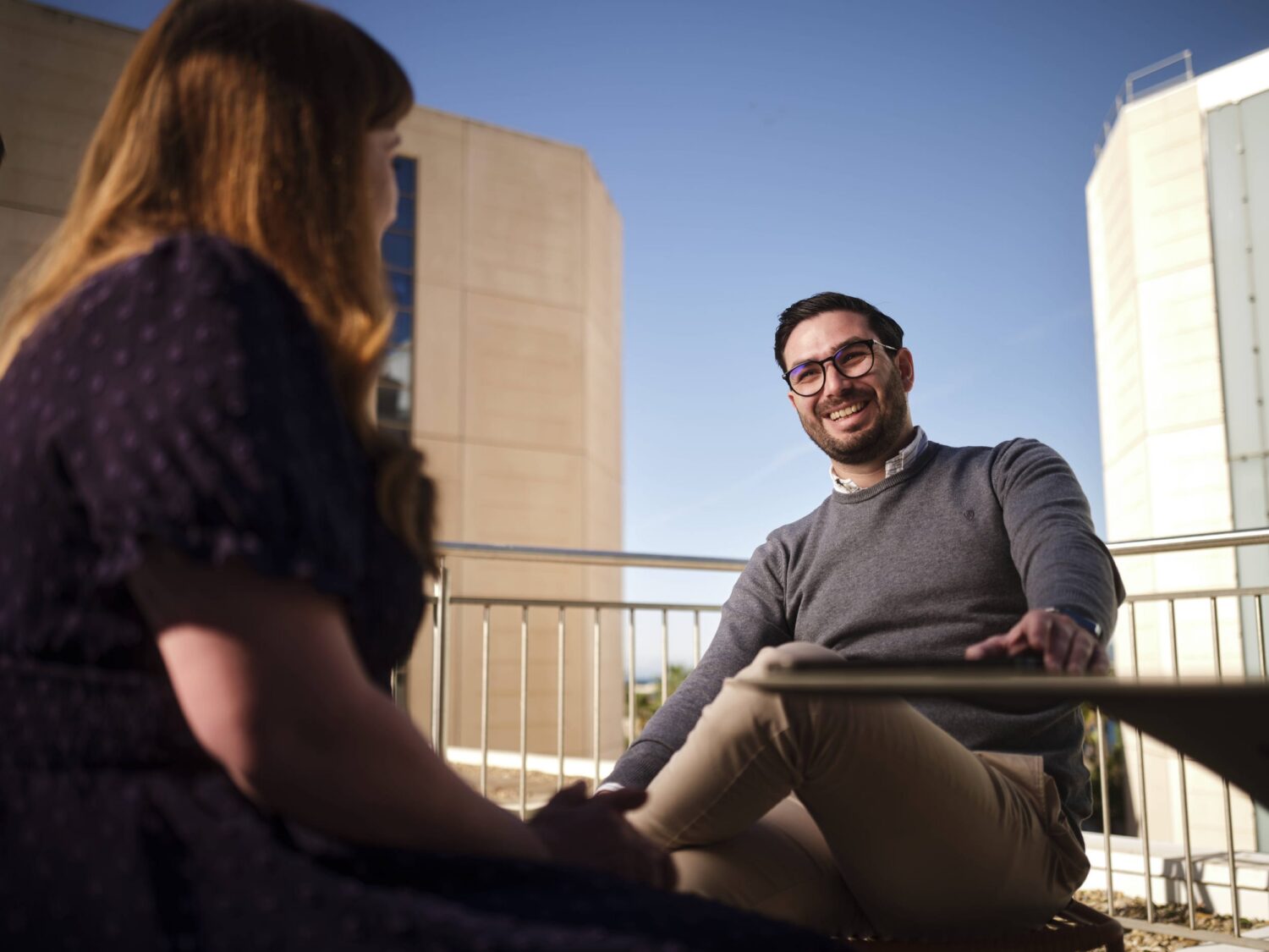 A man and a woman sitting on a rooftop balcony, talking with a clear blue sky and tall buildings in the background.