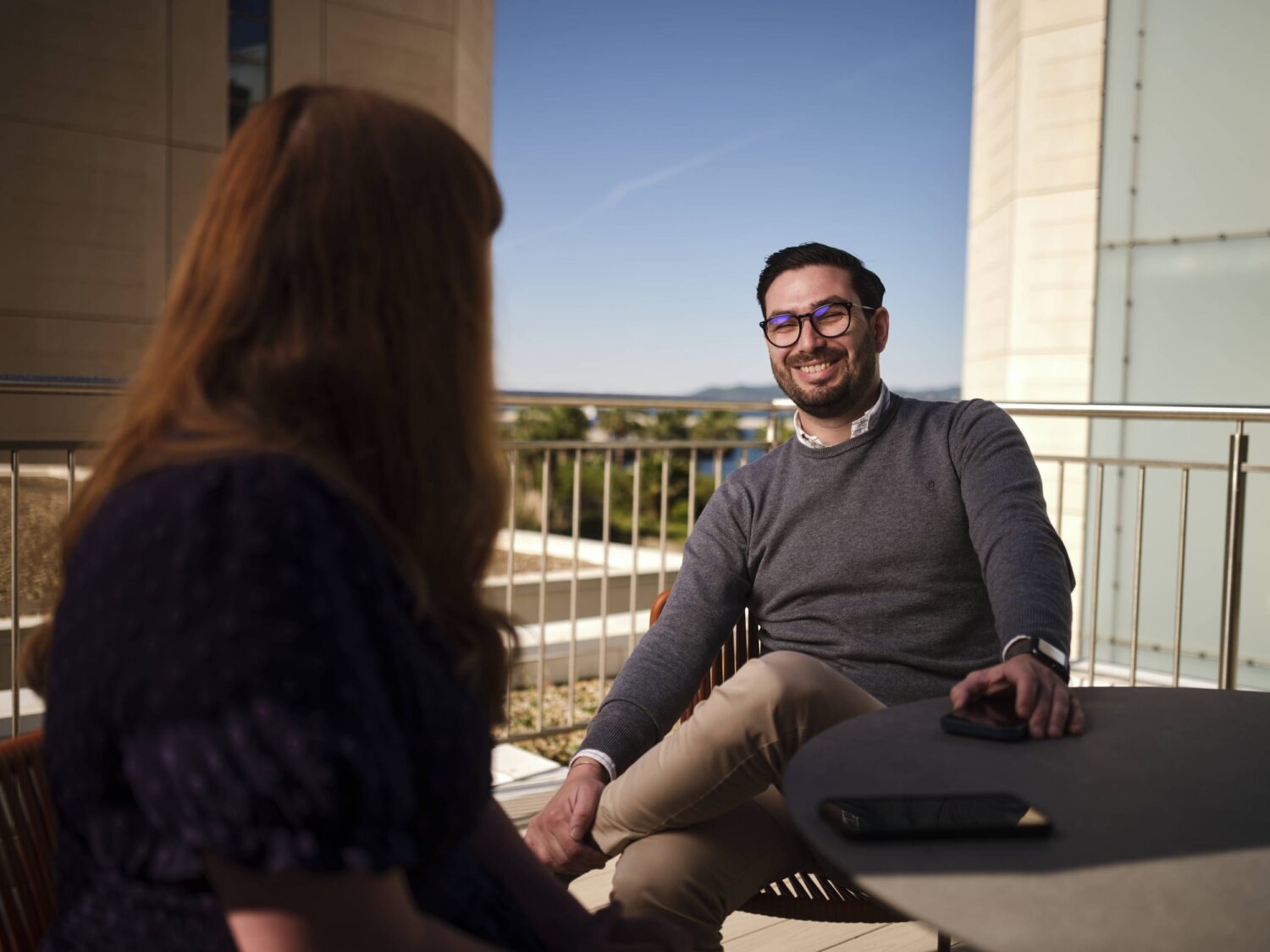 A man and a woman sit at an outdoor balcony table, smiling at each other with a laptop on the table and a clear blue sky in the background.