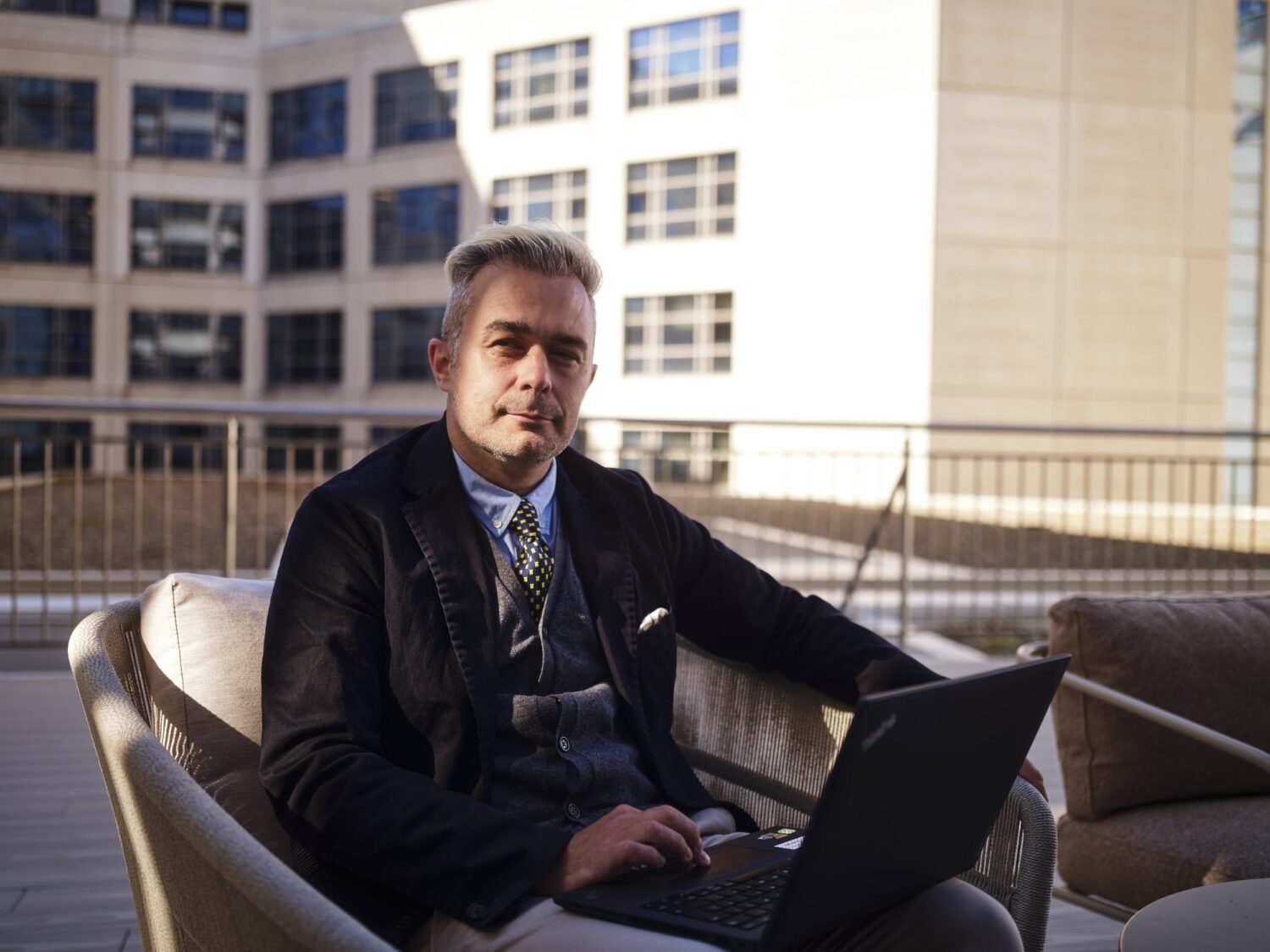 A man in a suit sits on an outdoor couch on a rooftop, using a laptop.
