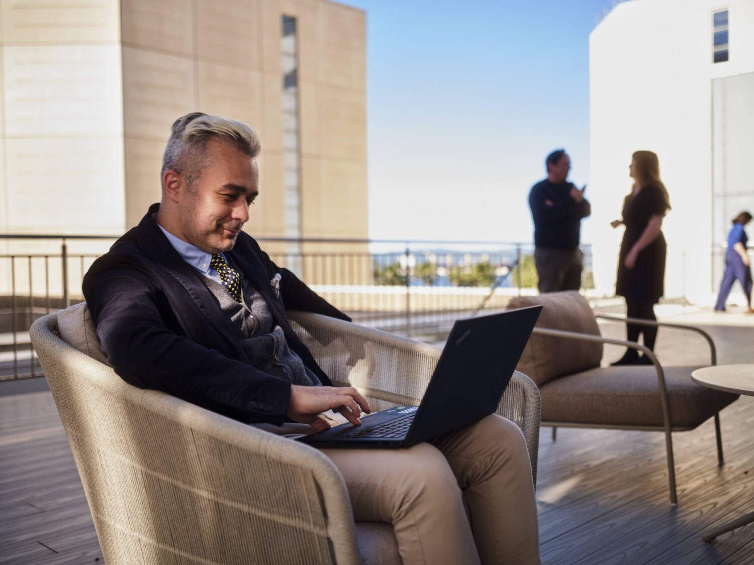 Man in a blazer sits on an outdoor patio chair using a laptop, with two people standing and chatting in the background.