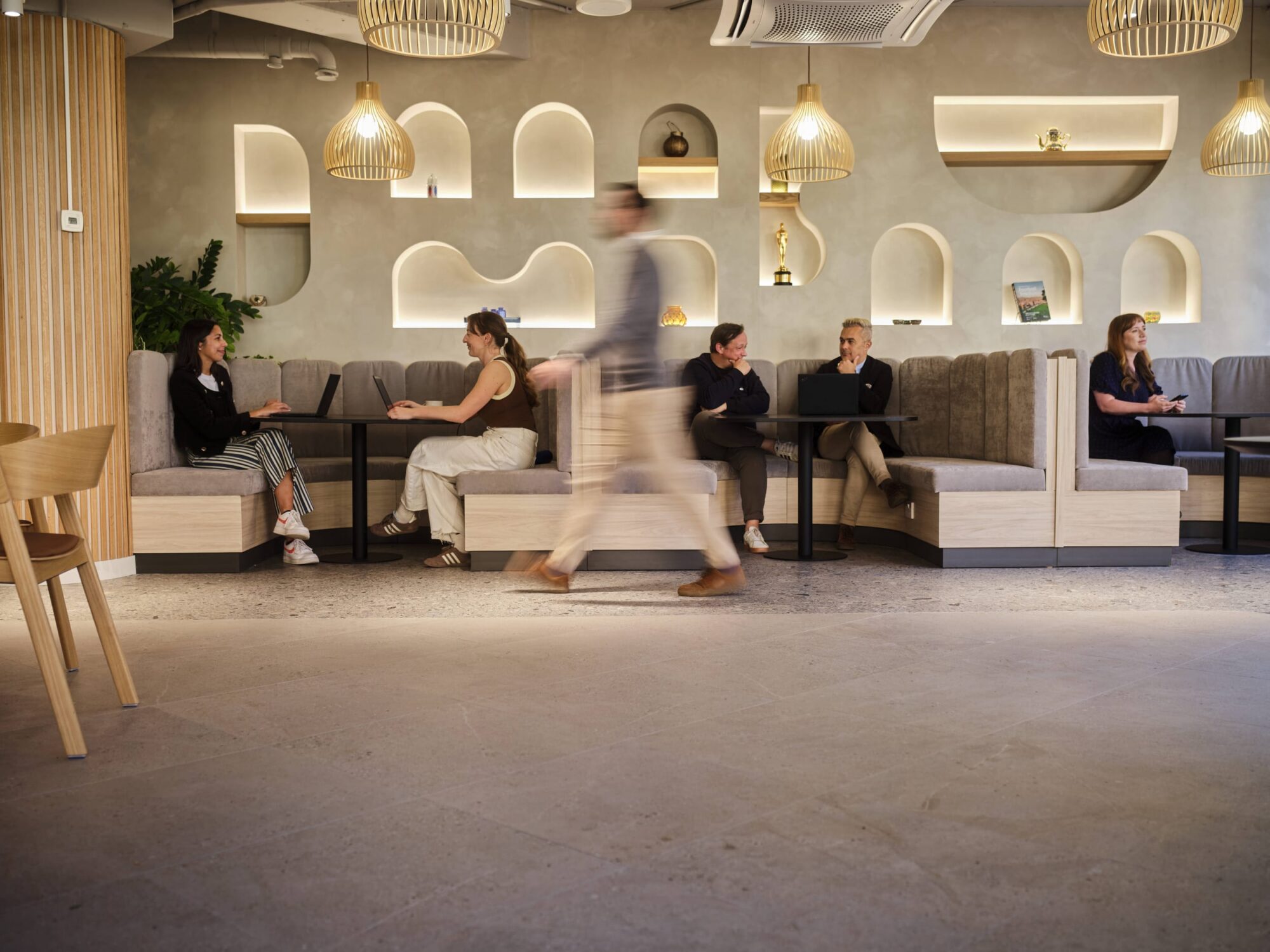 A blurred passerby walks past seated customers in a modern cafe with cushioned benches, arched wall niches, and warm hanging lights.
