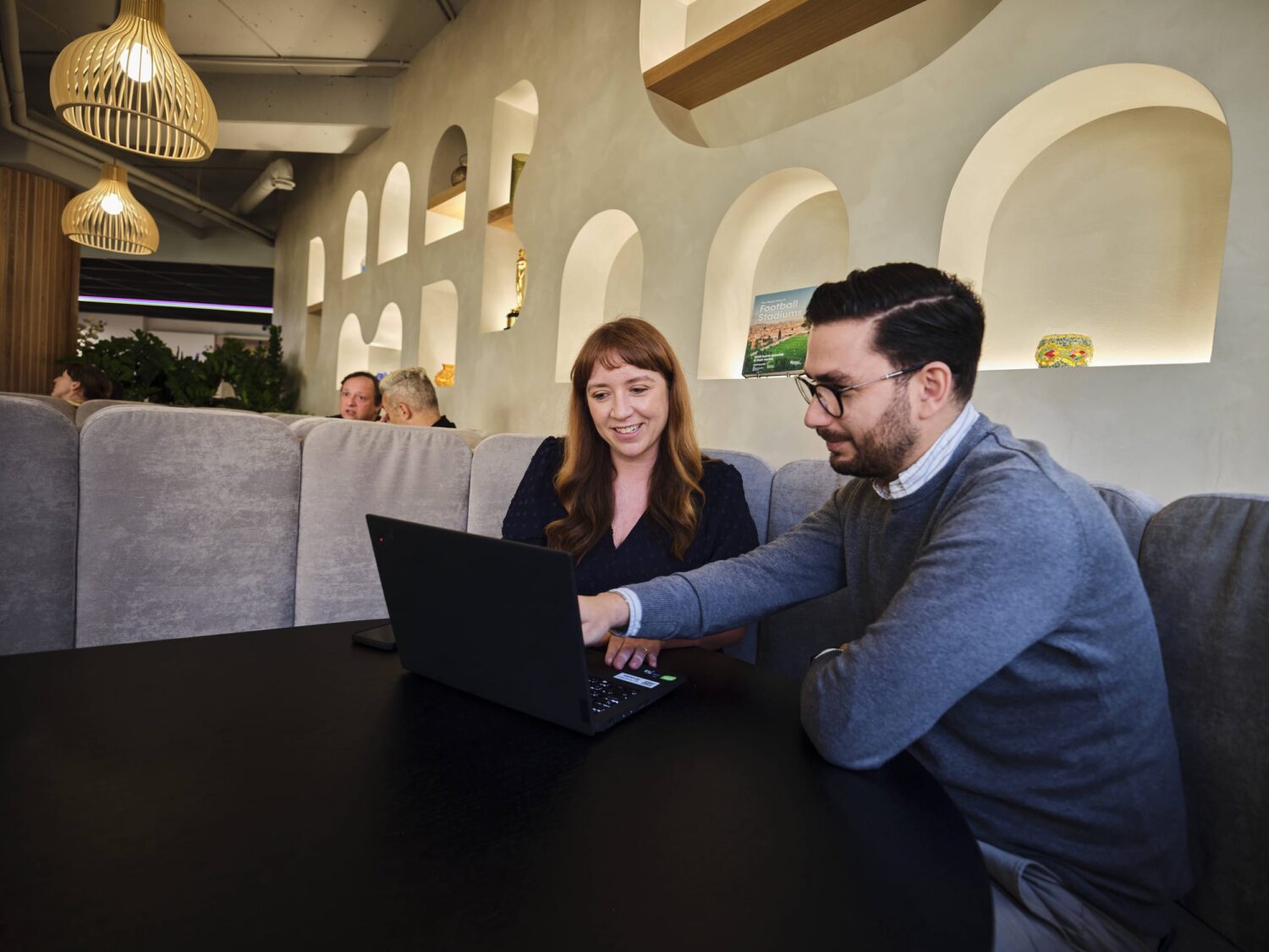 Two people sit at a table in a modern cafe, looking at a laptop together with arched wall niches and pendant lights in the background.