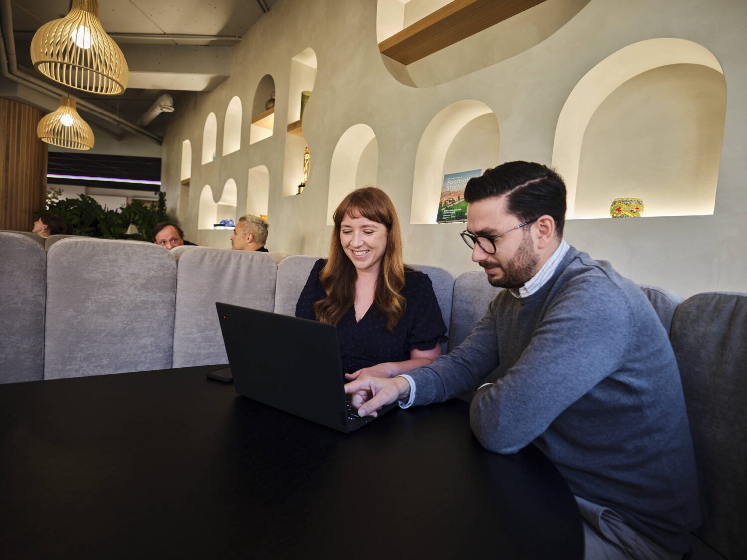 Two people sit at a dark table in a modern cafe, looking at a laptop together, with arched wall niches and hanging pendant lights in the background.