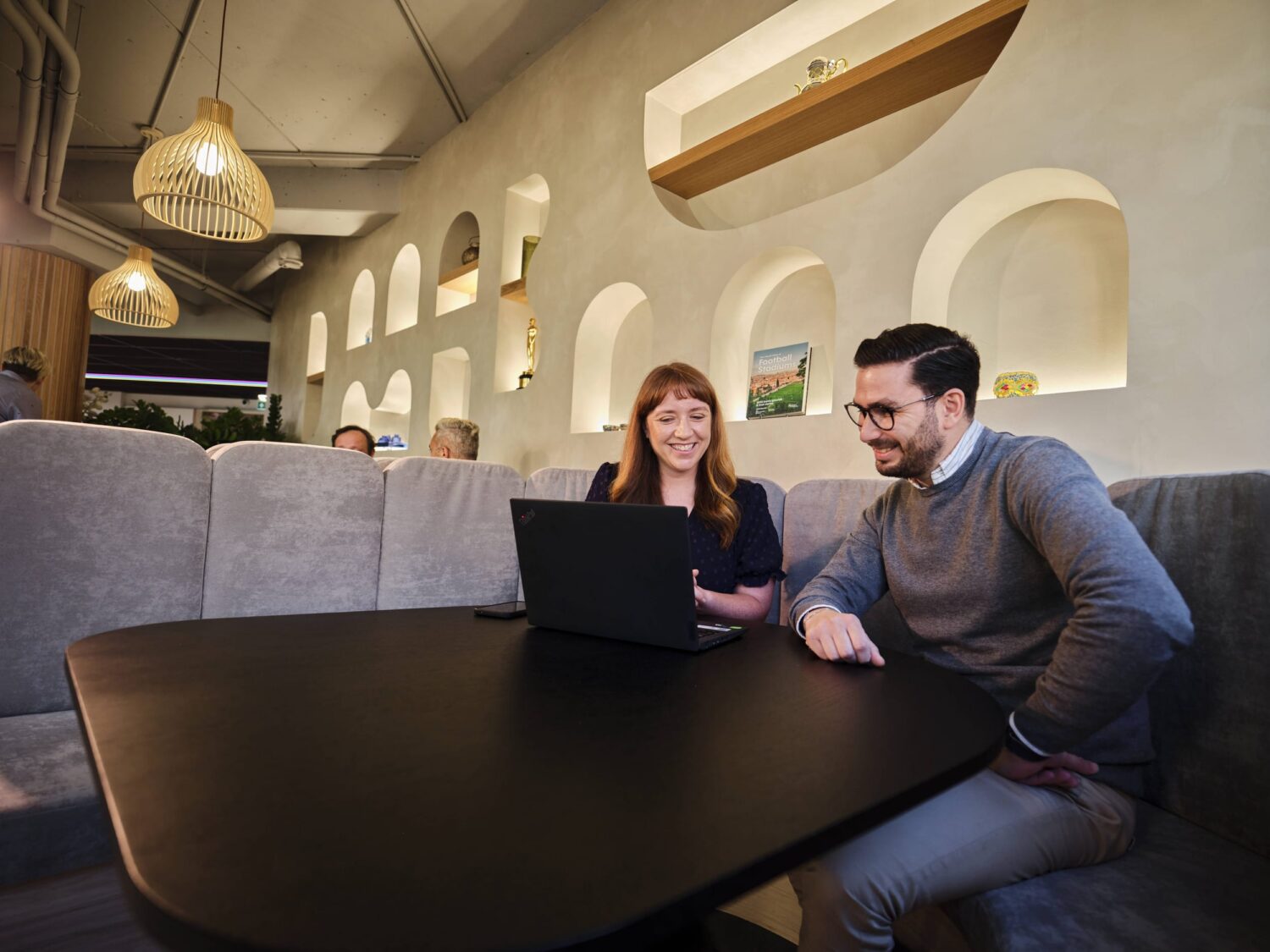 A man and woman sit on a gray couch at a dark wooden table in a modern cafe, looking at a laptop together.