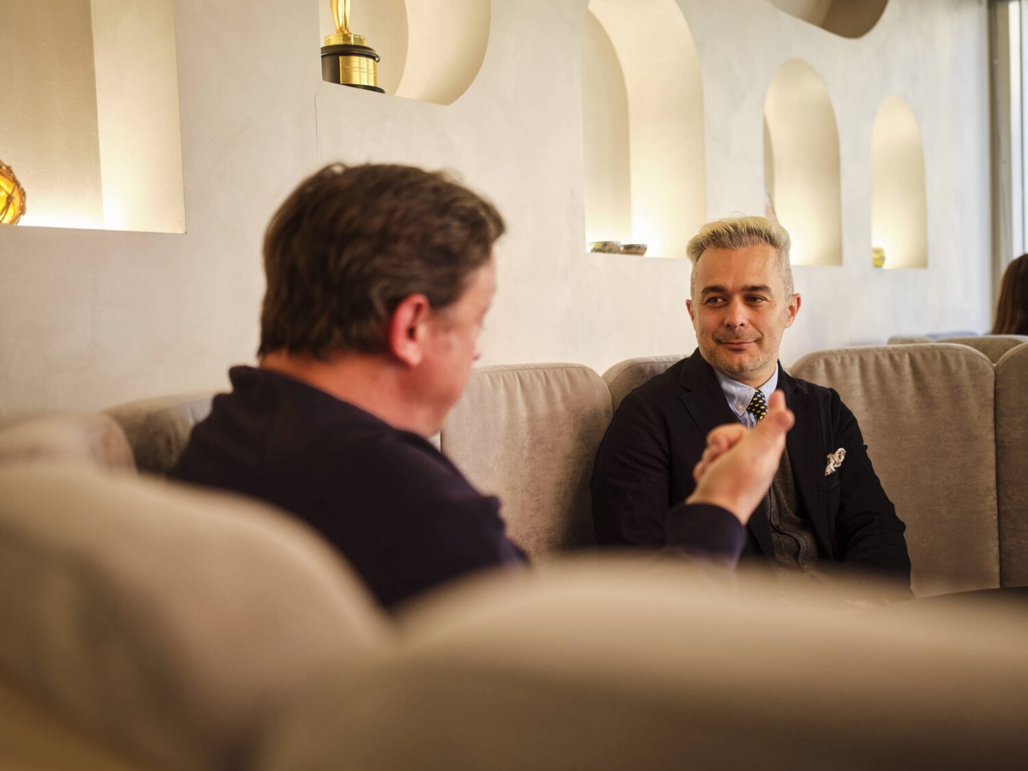Two men sit on a beige sofa in a modern lounge, chatting and holding drinks, with warm lighting and arched wall niches in the background.