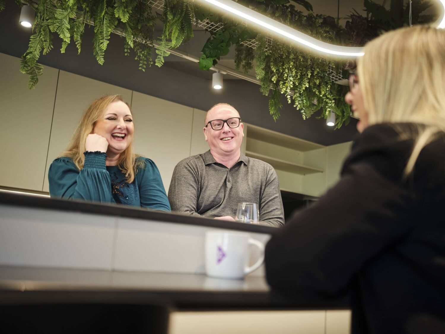 Two people sit behind a reception counter, smiling at a blonde person standing in front under greenery.