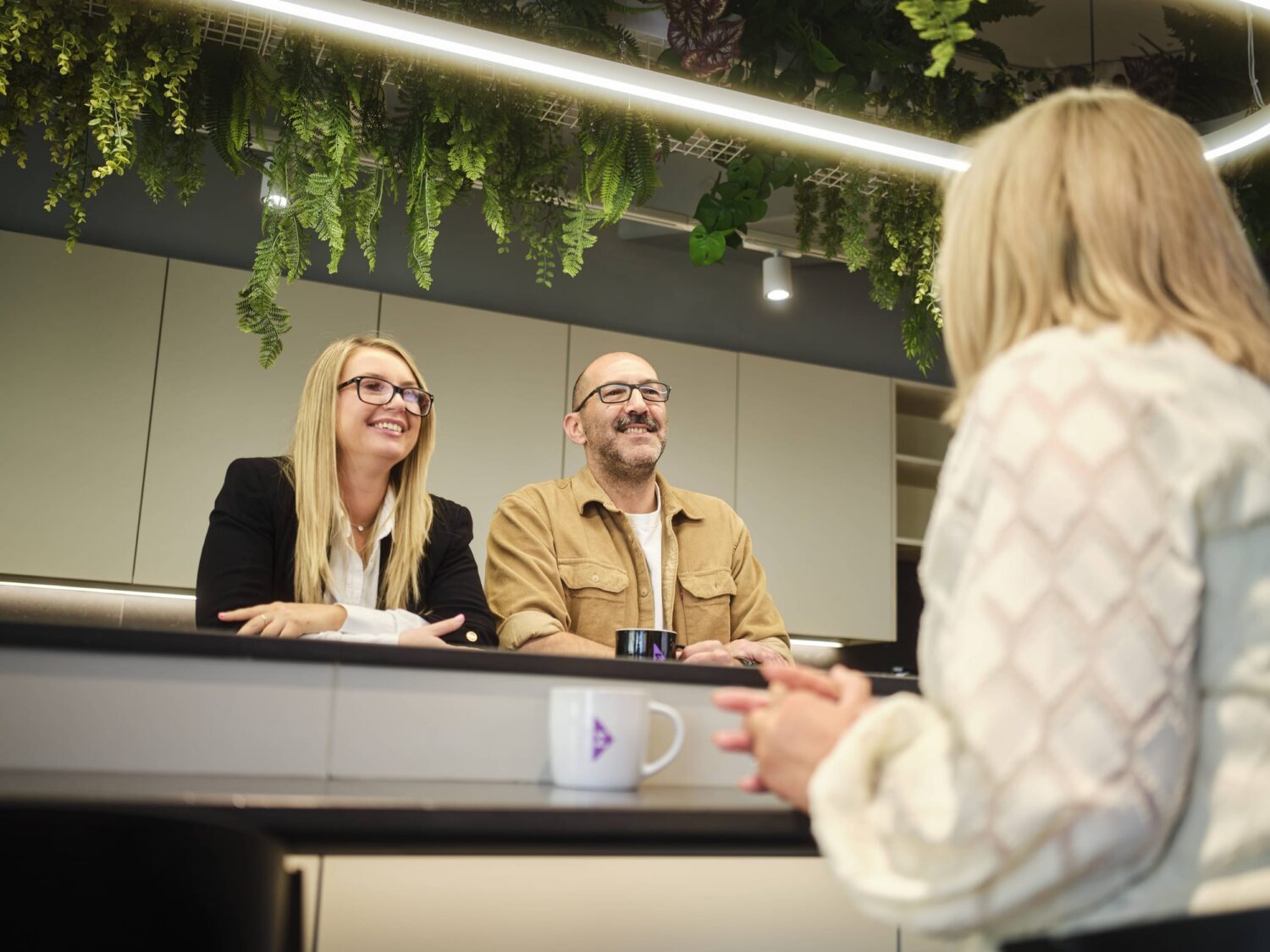 A customer with light-colored hair talks to two smiling staff members behind a counter in a modern, plant-decorated interior.