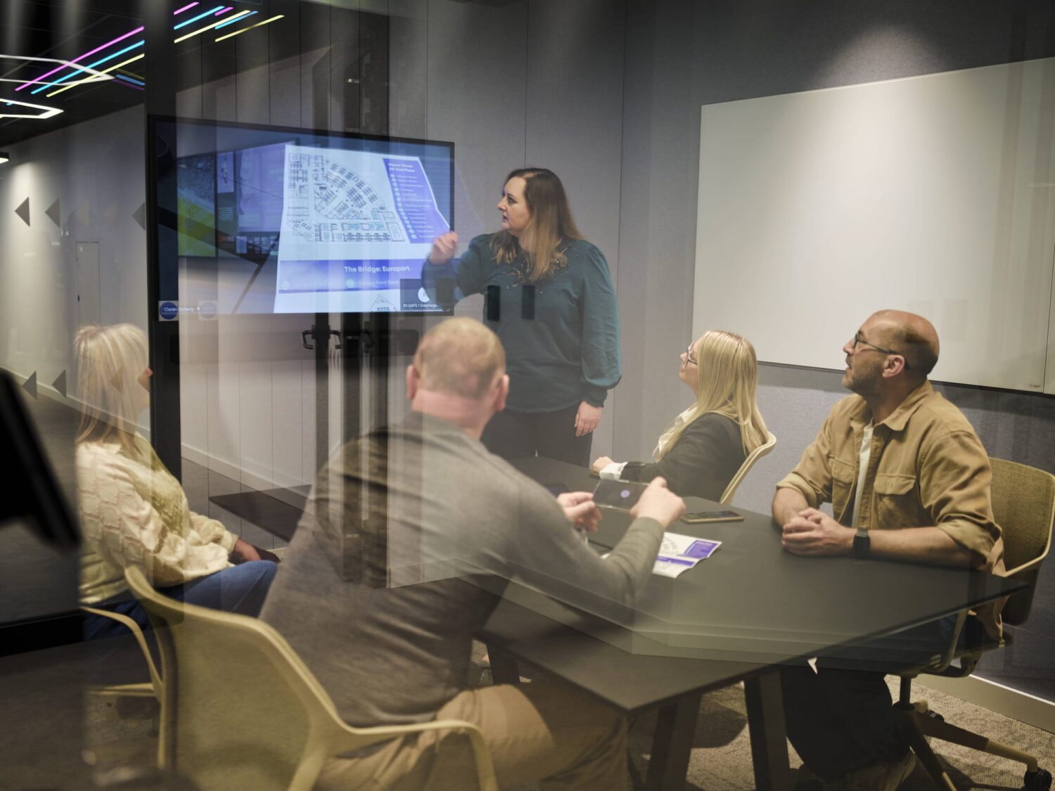 A presenter stands at the front of a conference room addressing a group of people seated around a table, with a projected diagram on the screen.