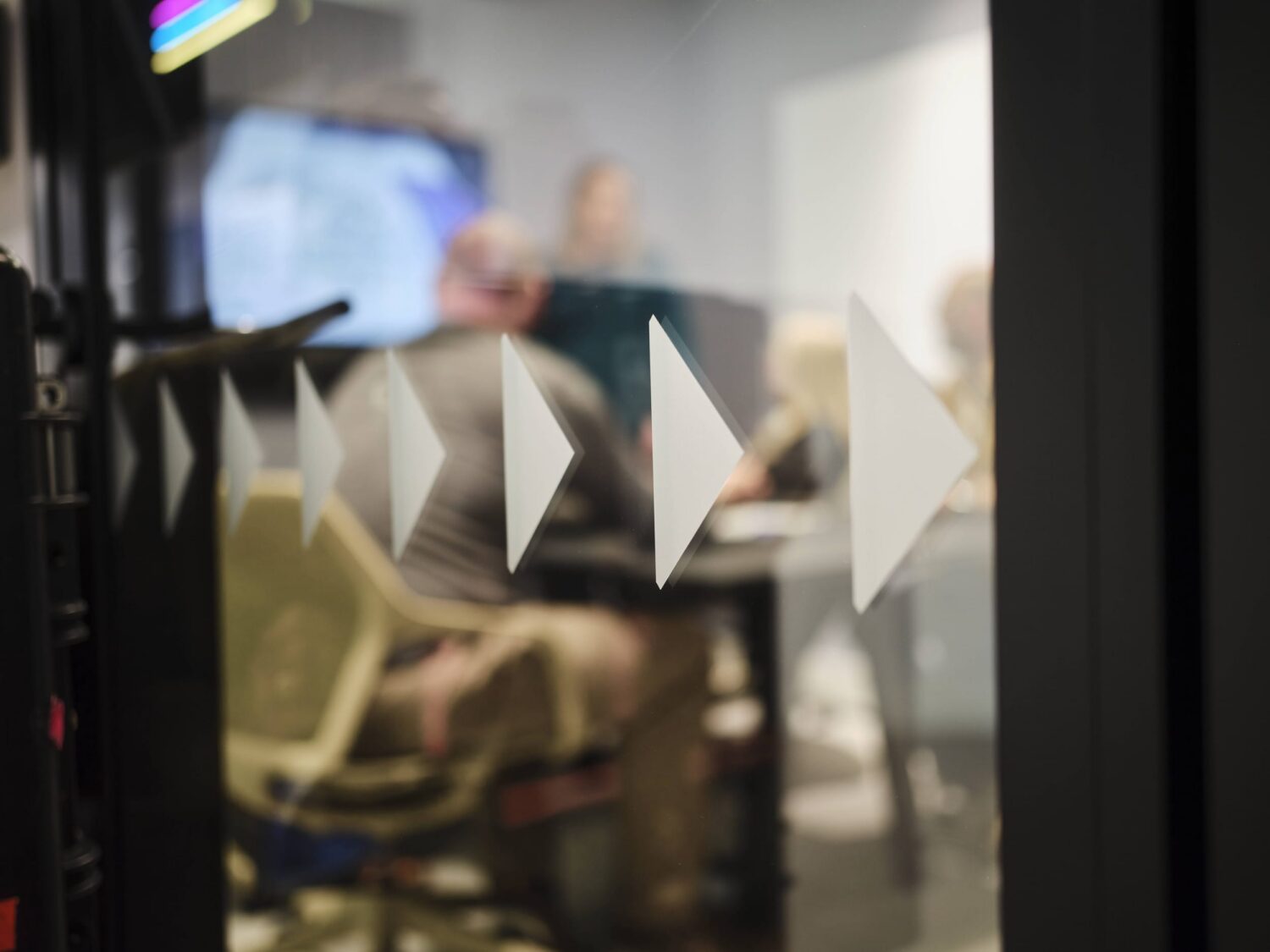 A glass partition with a row of white triangular shapes in the foreground and a blurred office meeting room in the background.