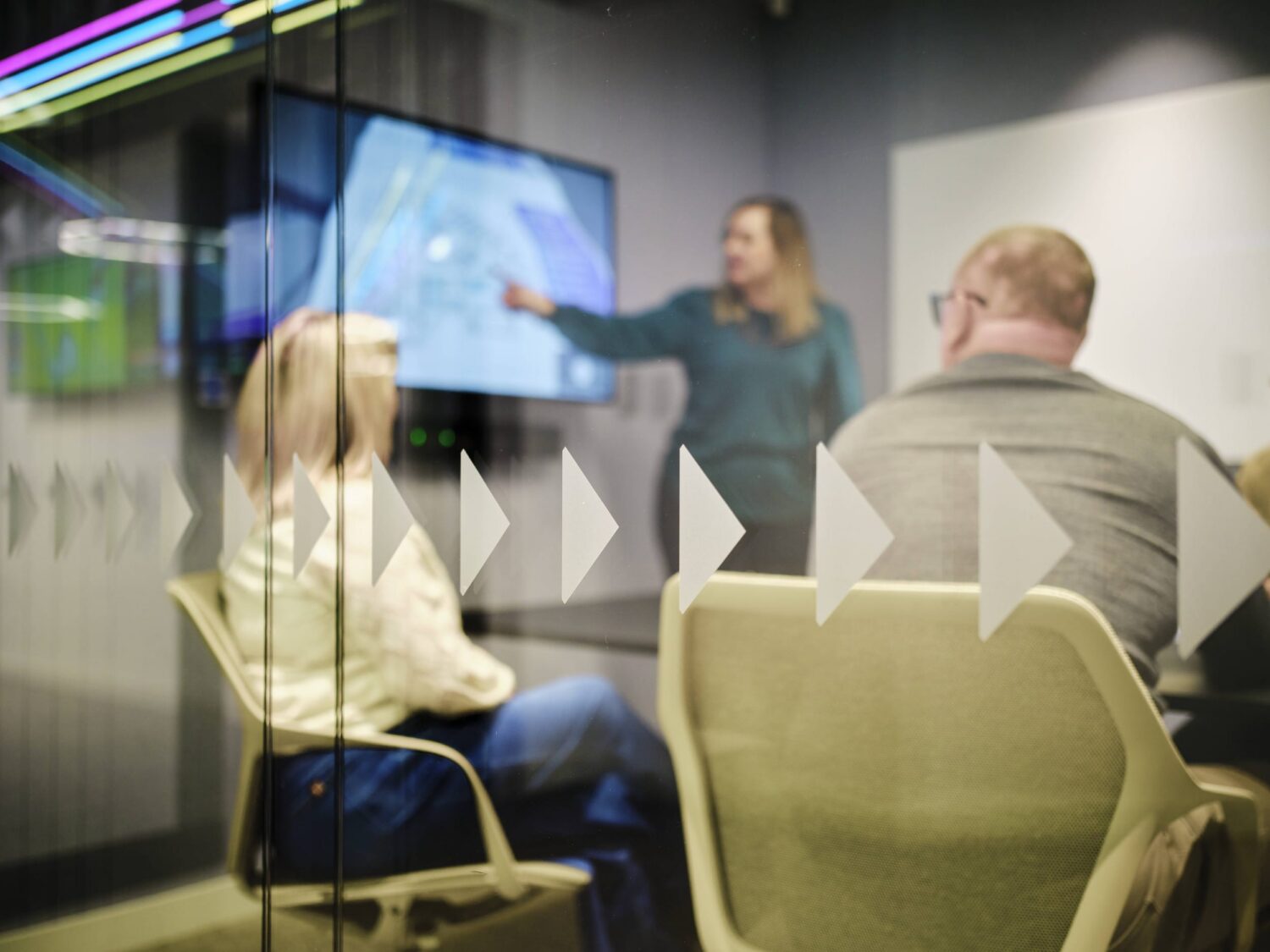 A presenter points at a large screen as seated colleagues watch in a modern conference room.