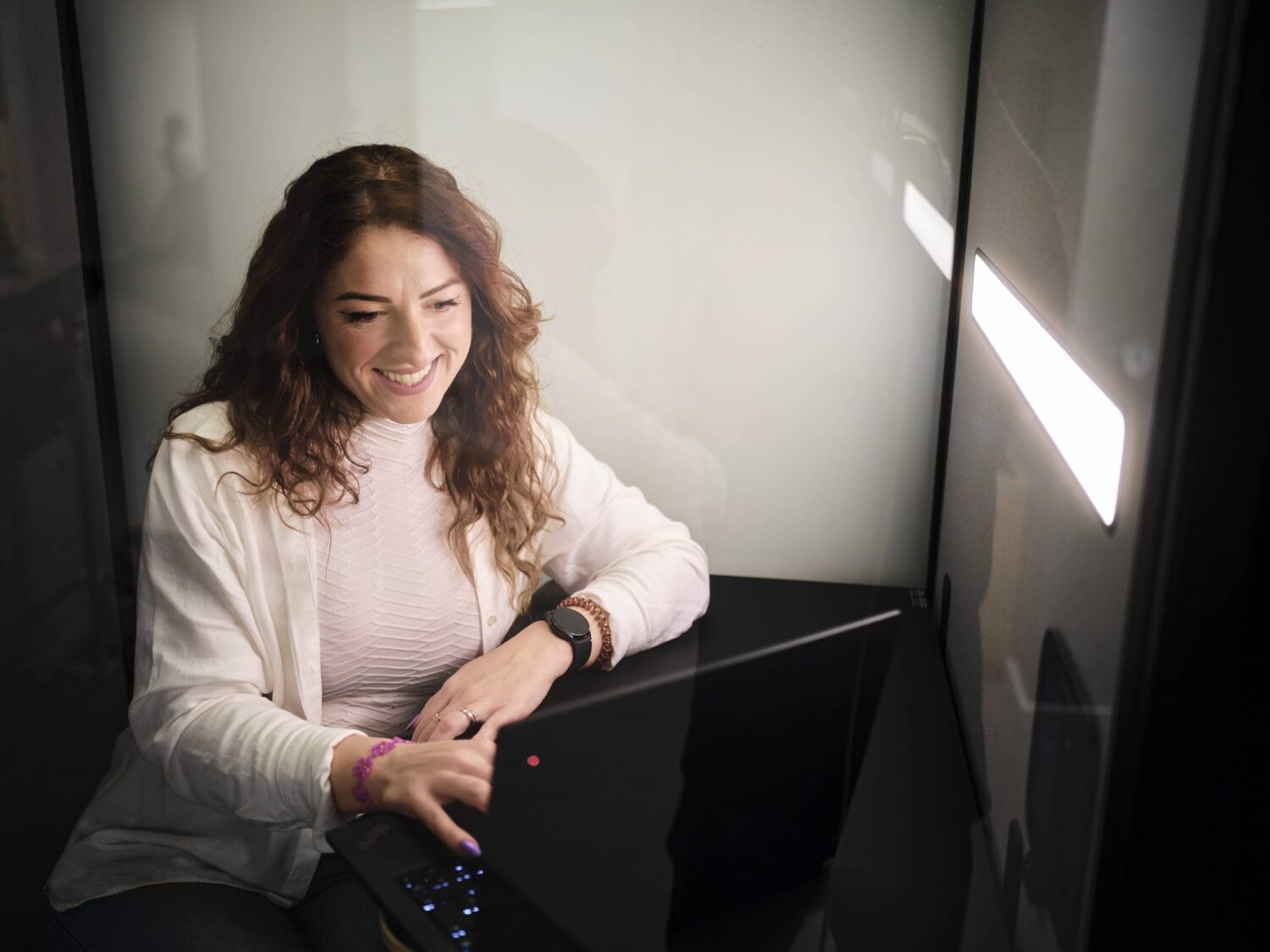 A smiling woman with curly hair sits in a private glass-walled booth using a laptop.