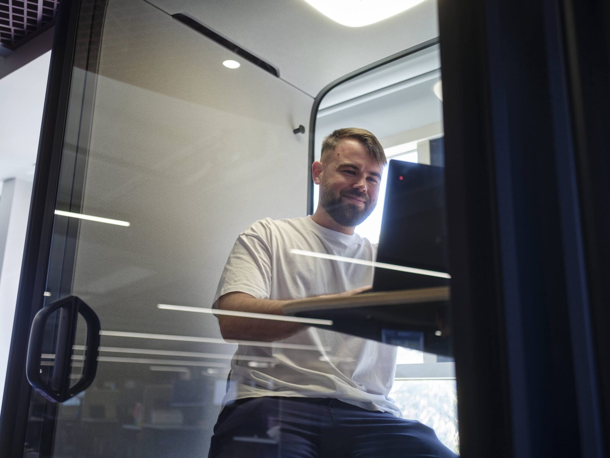 Bearded man in a white T-shirt stands in a glass-walled office, looking at a computer monitor.