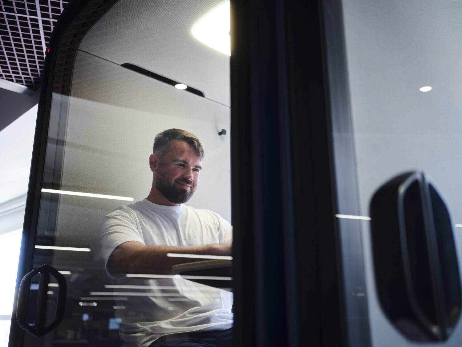 Bearded man in a white T-shirt sits inside a vehicle cab, looking out the window.