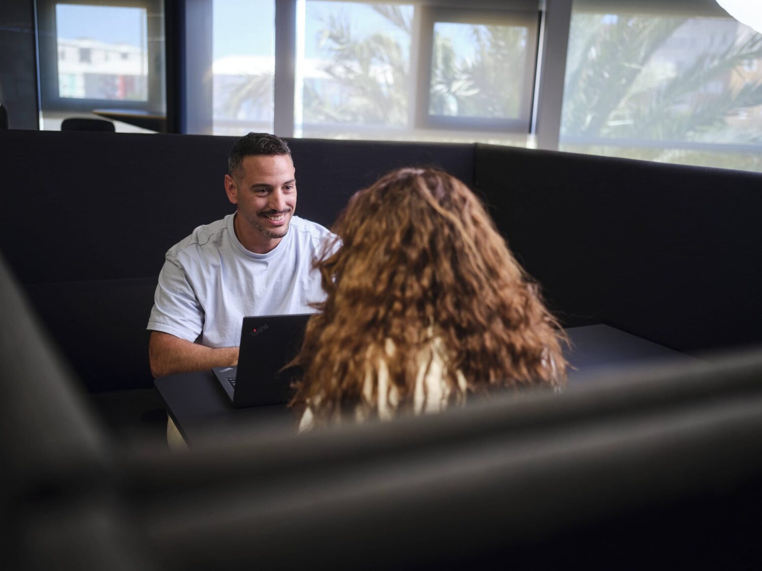 A man in a white T-shirt smiles while talking to a person with curly hair across a table in a bright indoor space.
