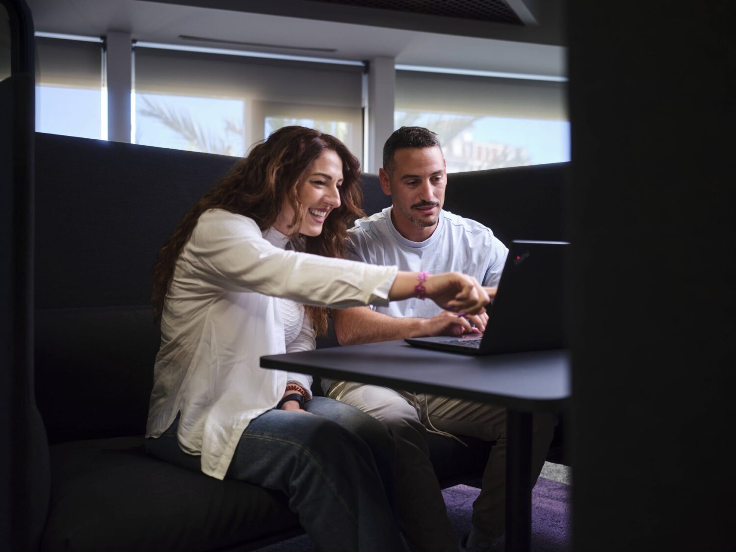 A man and a woman sit together at a small table, looking at a laptop with the woman pointing at the screen.