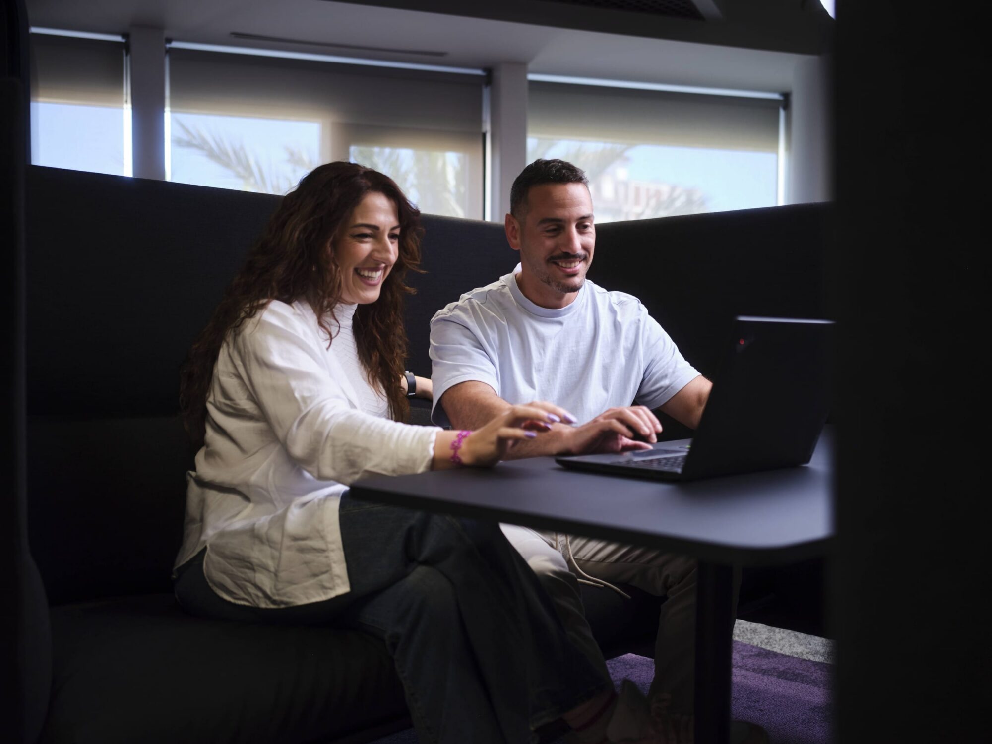 Two coworkers sit in a cafe booth, smiling while working together on a laptop.