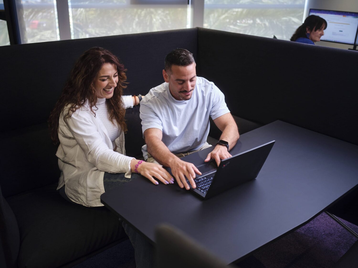 Two people sit at a dark table, working on a laptop in a modern office lounge.